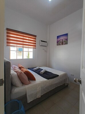 Een enkel bed met een witte matras, oranje en roze kussens, een blauwe deken en een witte handdoek. Er is een raam met oranje luiken, een wandairconditioner en een ingelijst schilderij aan de muur.