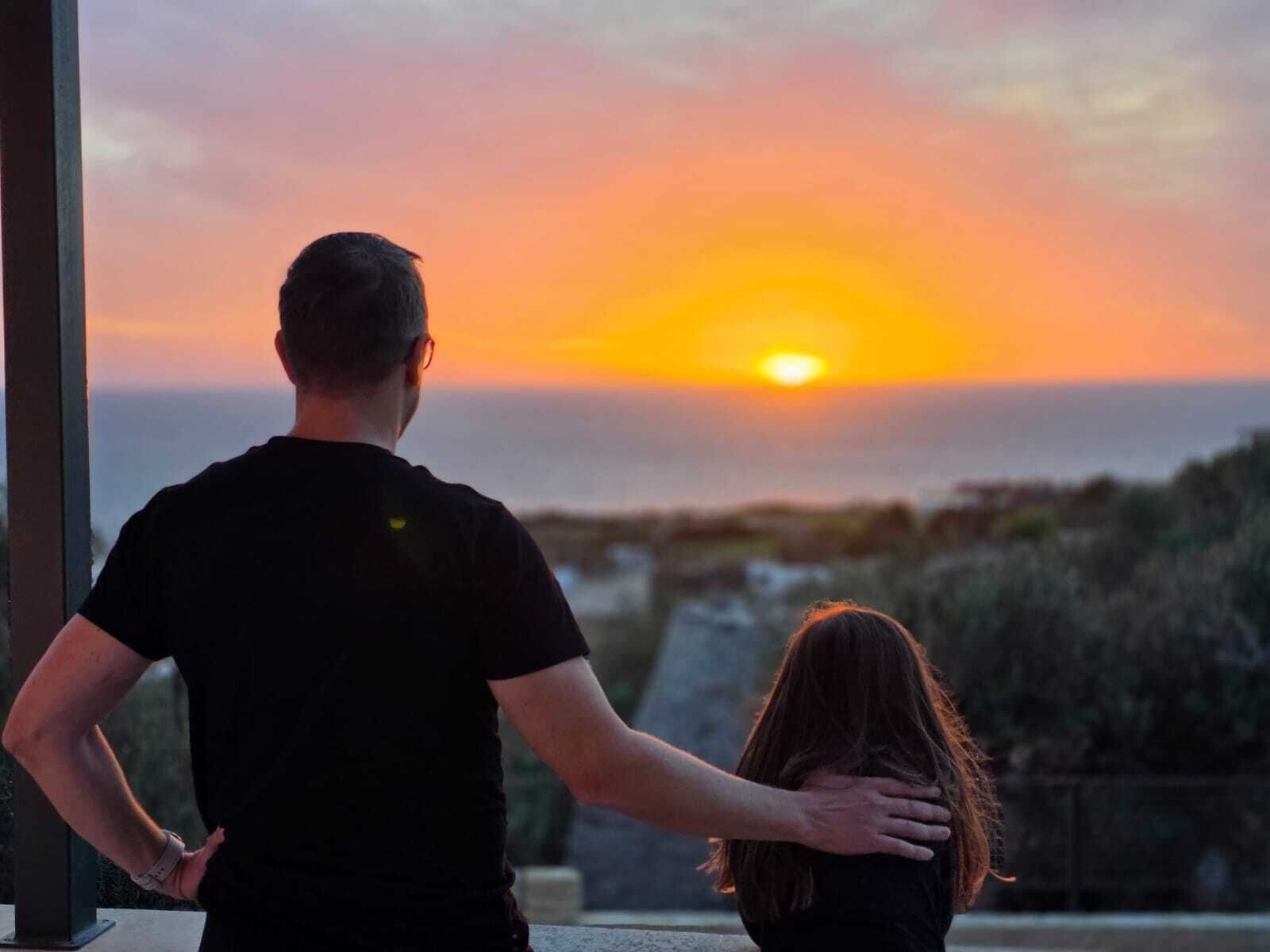 Un hombre y una mujer de pie junto a una ventana, contemplando un atardecer con vistas al mar y colinas.