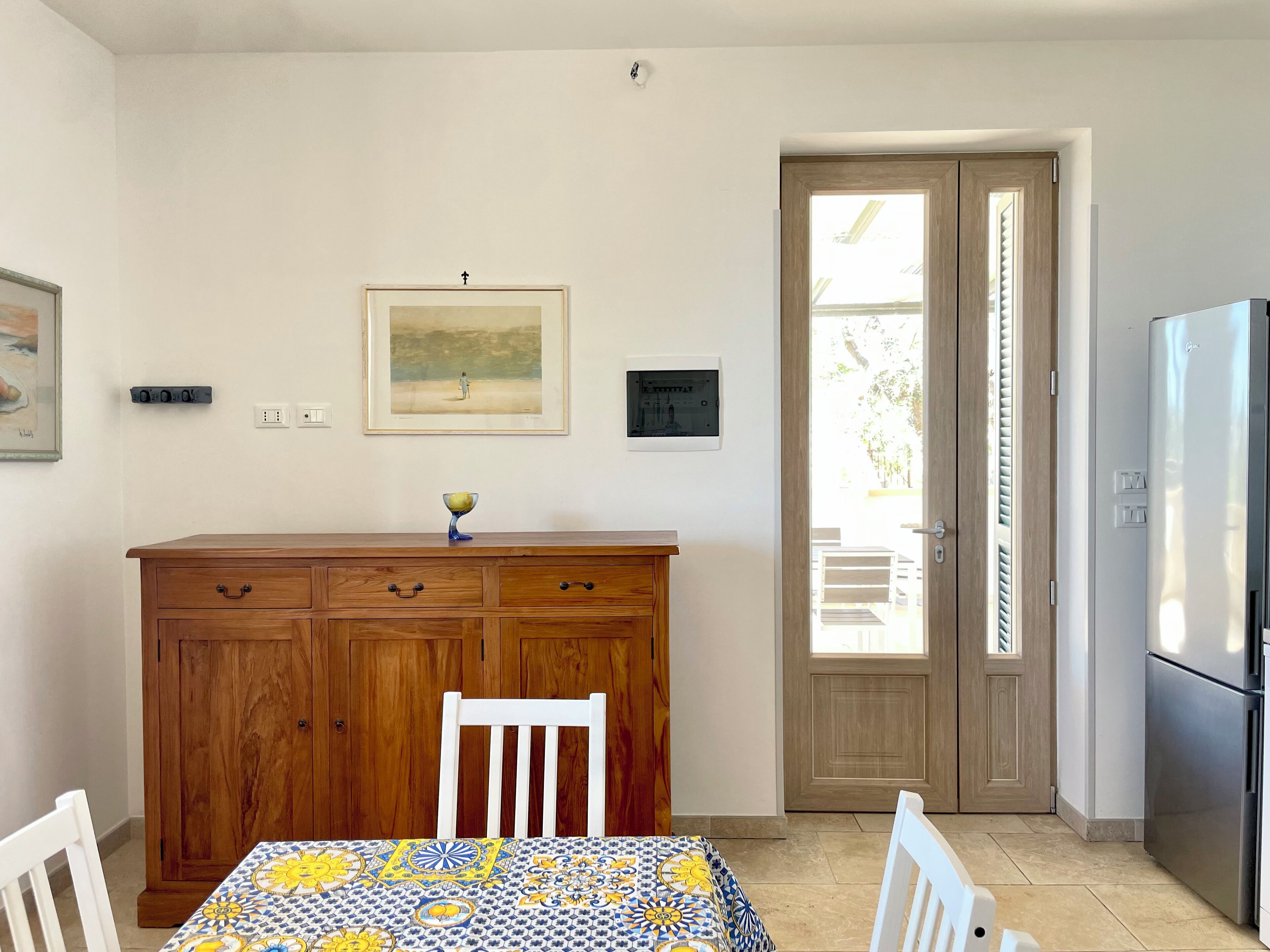 A kitchen with a wooden cabinet, a dining table with a patterned tablecloth, a refrigerator, and a door leading outside.