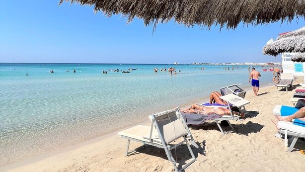 Una spiaggia con acque cristalline turchesi, lettini e un ombrellone di paglia che offre ombra.
