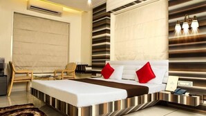 A modern hotel room with a large bed, a desk, and a chair. There is a large window with blinds, a wall-mounted air conditioner, and a bedside table with a lamp. - Hotel Fun City (Navsari)