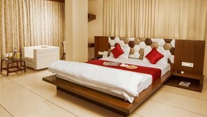 A modern hotel room with a large bed, a bedside table, a small table with a chair, and a large window with curtains. - Hotel Fun City (Navsari)