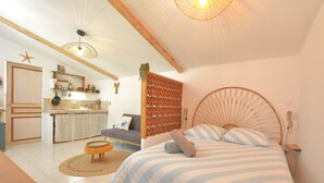 Une chambre à coucher avec un lit, une table de chevet et une commode. Il y a une fenêtre avec des rideaux, un plafond en bois et une étagère fixée au mur, sur laquelle sont posés divers objets.