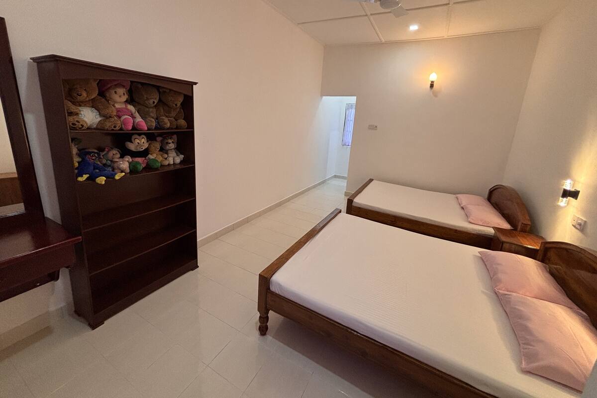 A bedroom with two single beds, a wooden shelf with stuffed toys, a ceiling fan, and a wall-mounted light.