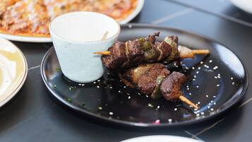 A plate of skewered meat with a dipping sauce and a pizza in the background.