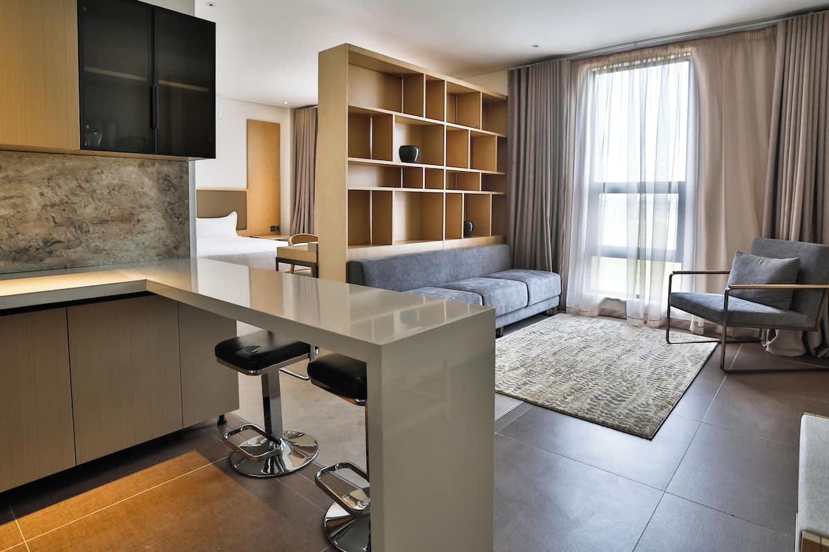 Una habitación de hotel moderna con kitchenette, un sofá, una silla y un ventanal con cortinas.