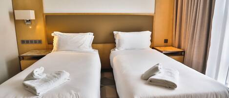 A hotel room with two beds, each with white linens and towels, a headboard, bedside lamps, and a nightstand.