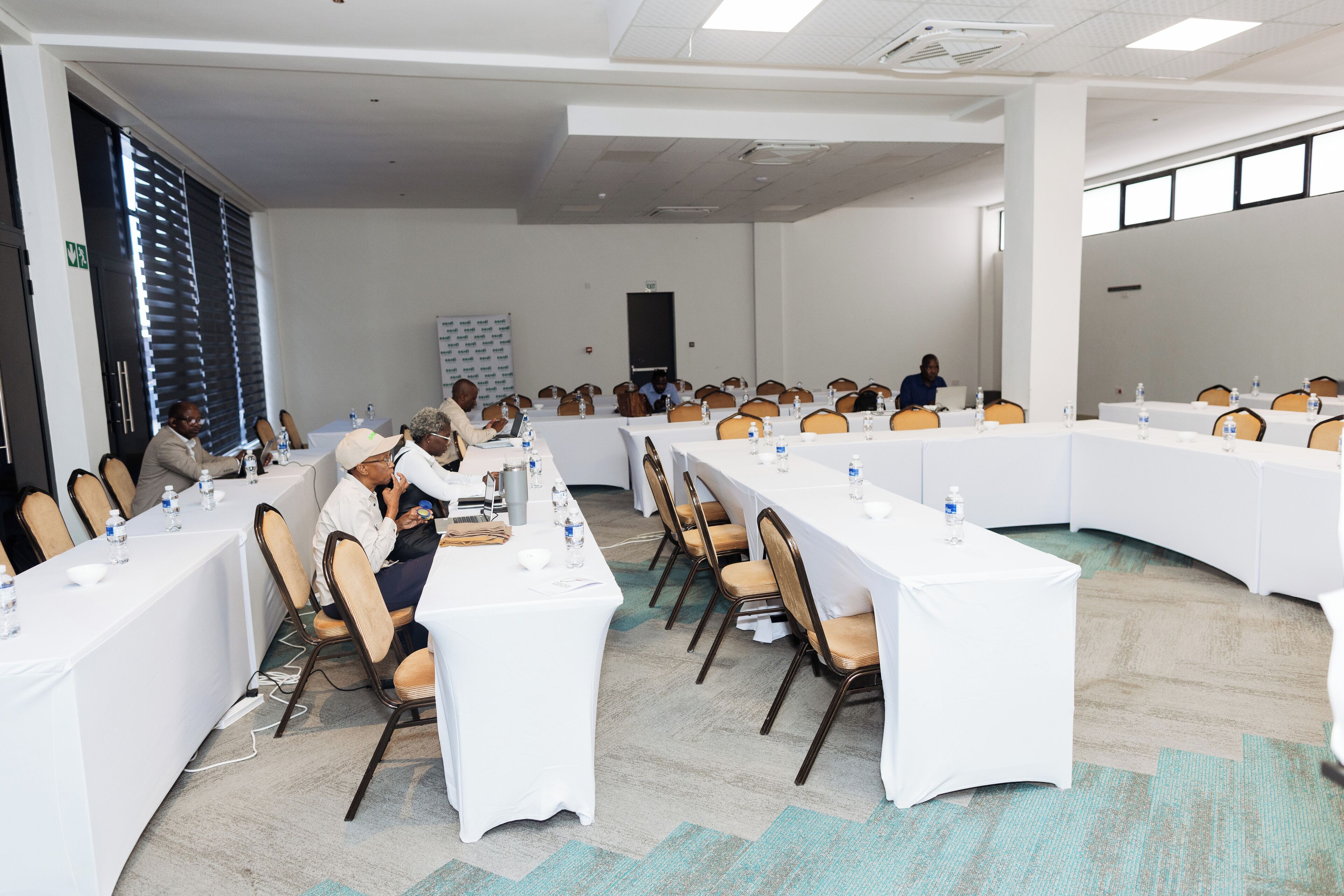 Un salón de conferencias con mesas largas, sillas y botellas de agua.