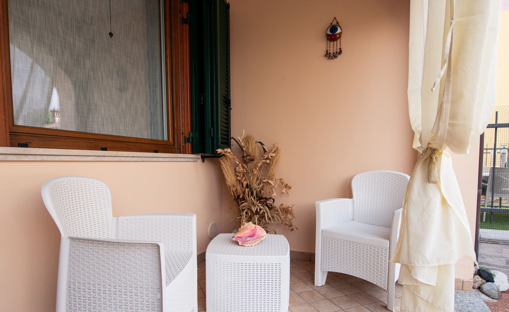 A patio with two white wicker chairs, a small table with a pink flower, and a decorative vase with dried plants.