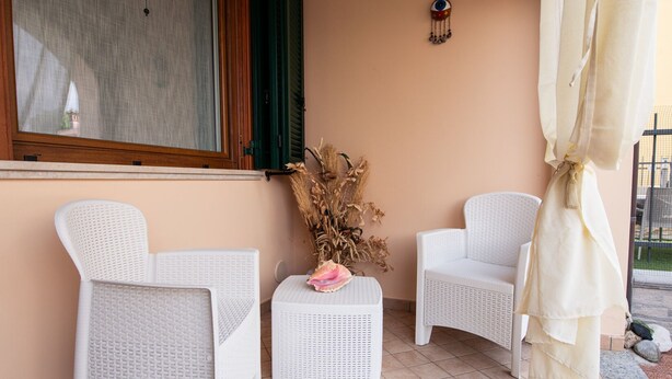 A patio with two white wicker chairs, a small table with a pink flower, and a decorative vase with dried plants.