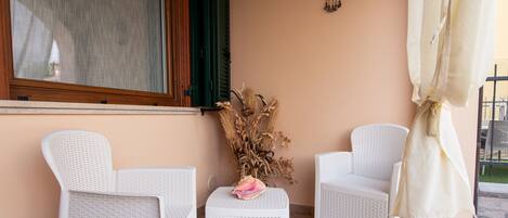 A patio with two white wicker chairs, a small table with a pink flower, and a decorative vase with dried plants.