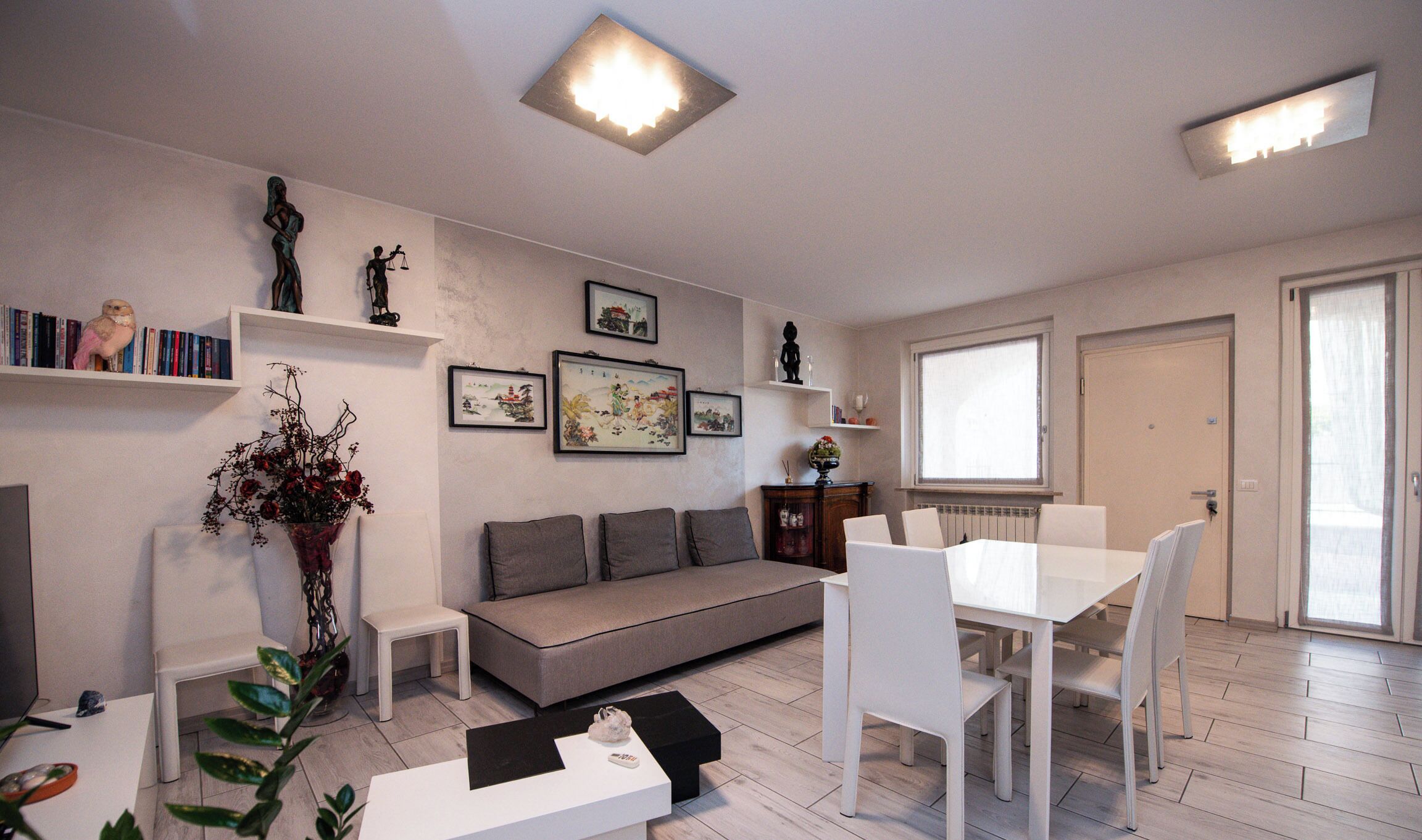 A modern living room with a sofa, dining table, and chairs. There are bookshelves with books and decorative items, a fireplace, and a large window.