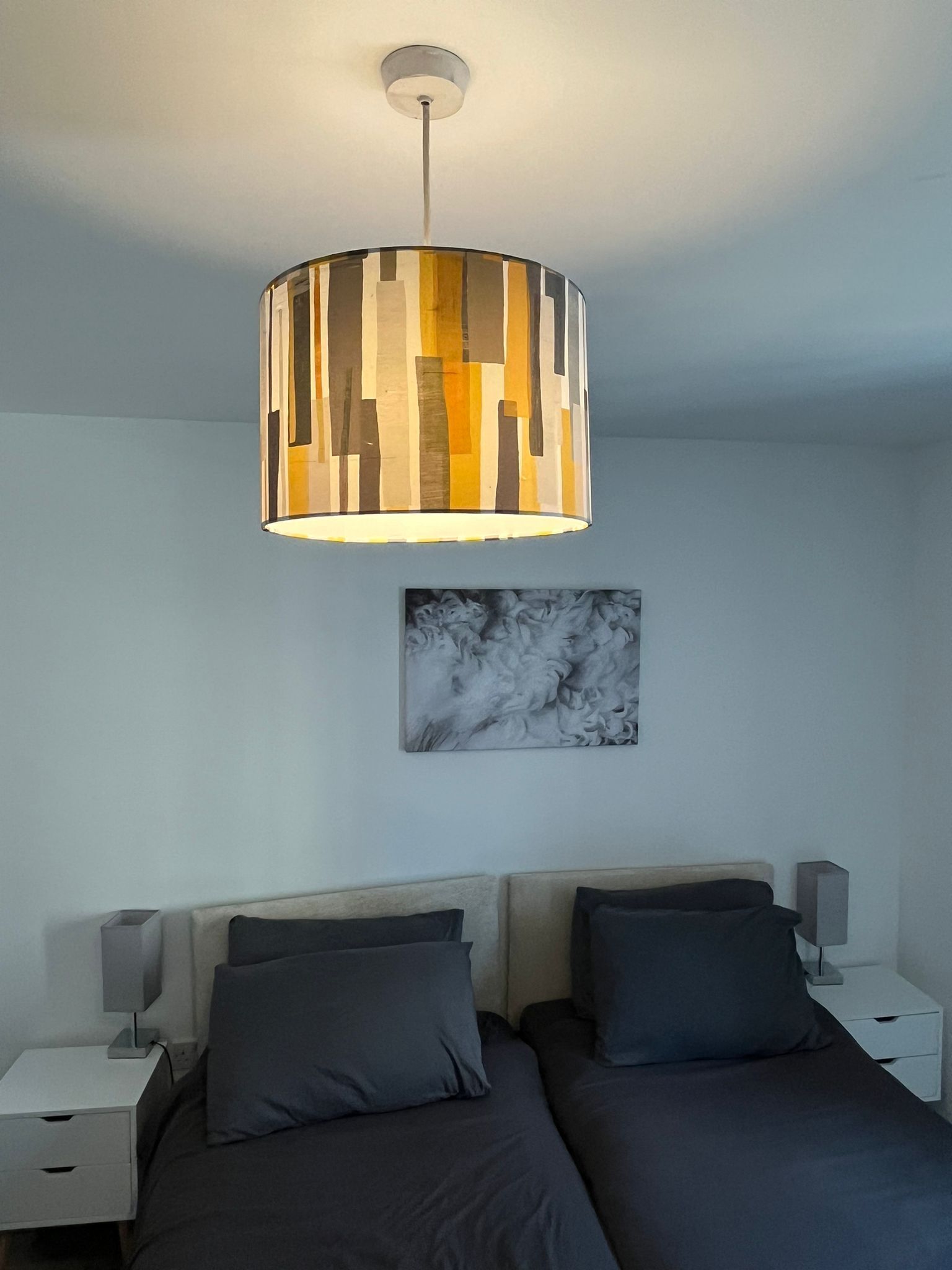 Ett sovrum med en modern säng, två sängbord, en abstrakt tavla som hänger på väggen och en unik, cylinderformad hänglampa.
