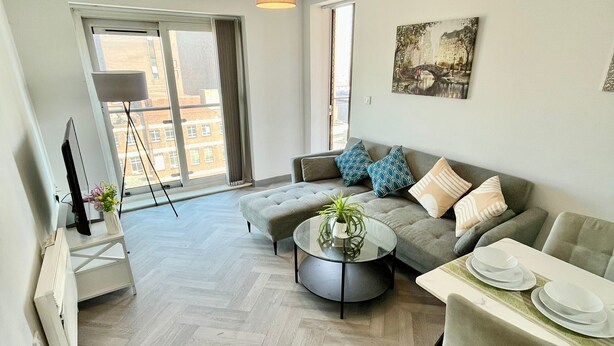 A modern living room with a sofa, coffee table, and dining table. There is a large window with a view of buildings. - Cosy Apartment in the City Centre of Birmingham, walk to everything (Birmingham)
