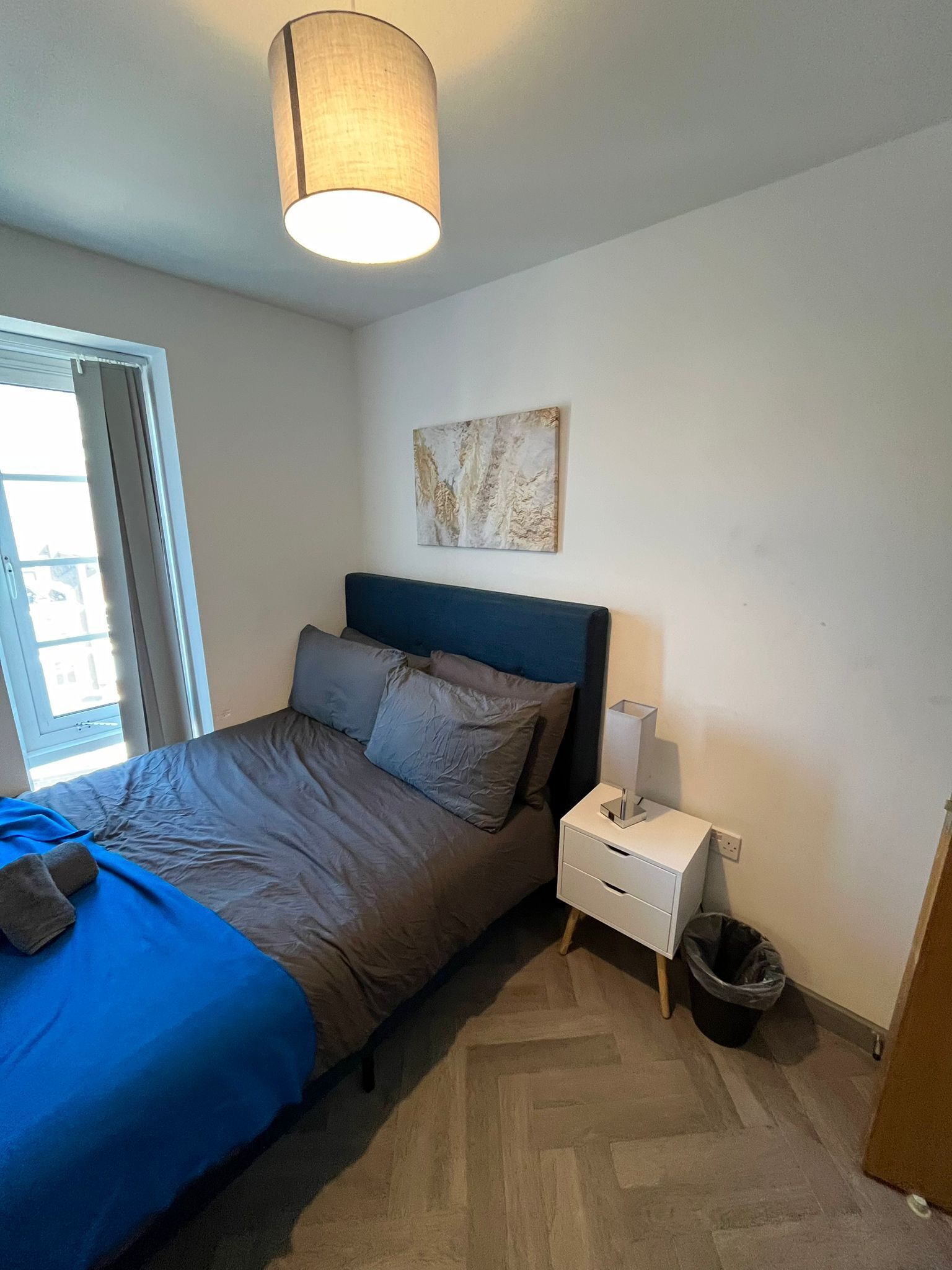 Une chambre à coucher avec un lit, une table de chevet, une lampe, une poubelle et une fenêtre avec des rideaux.
