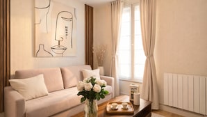 A living room with a sofa, a coffee table with a vase of flowers, a painting on the wall, and a window with curtains. - Courtois (Paris)