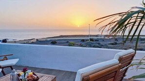 Una terraza con vistas a la puesta de sol sobre el mar, amueblada con sillas de madera y una mesa con alimentos para el desayuno.