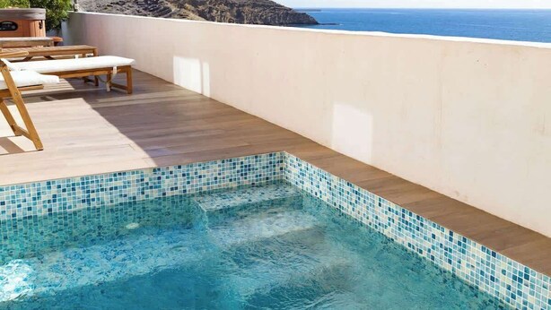 Una piscina con borde de azulejos, plataforma de madera y sillas de descanso.