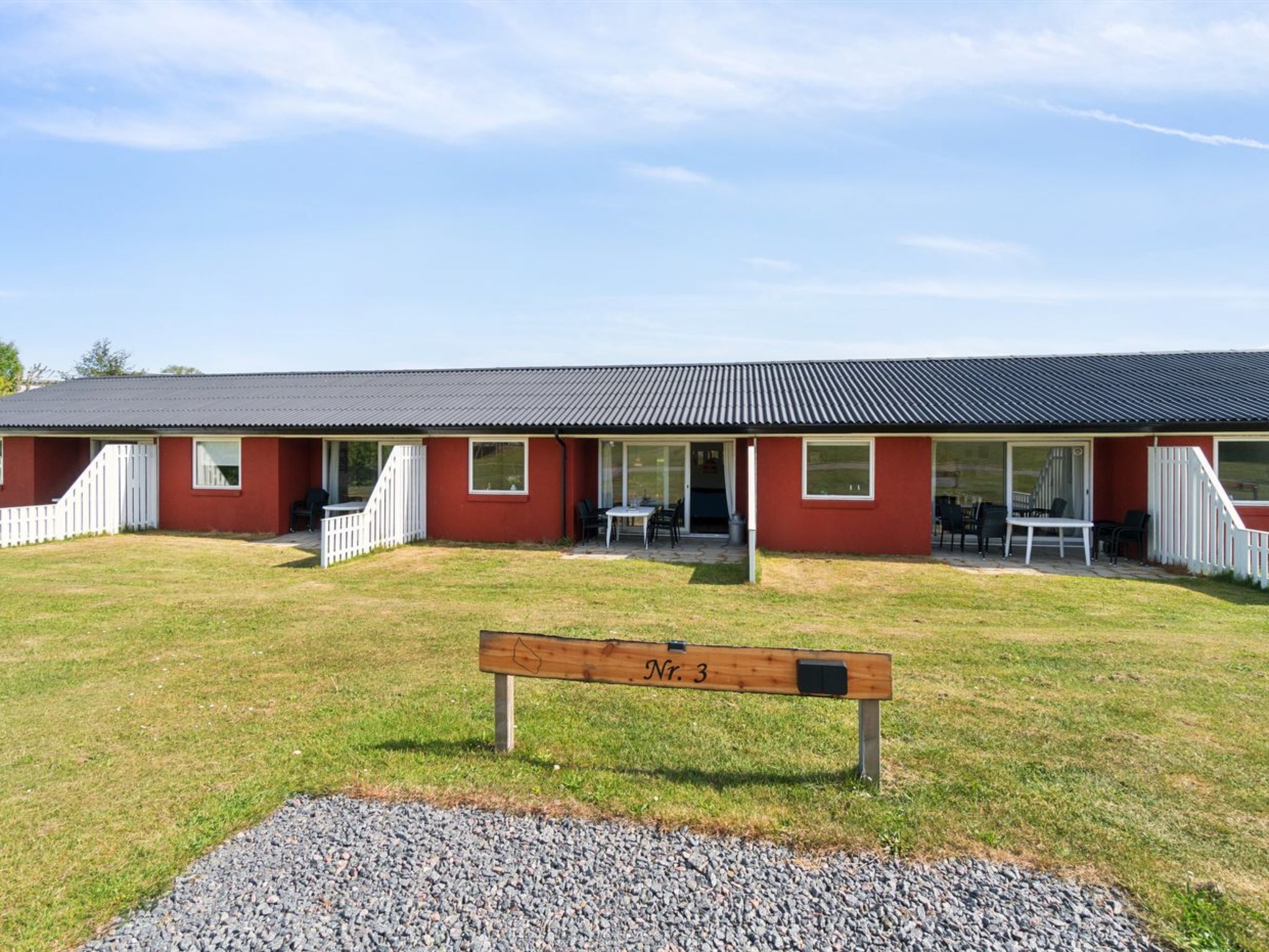A red building with a covered patio area, a gravel path, and a wooden bench labeled "Nr. 3".