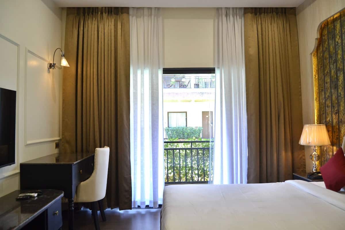 Habitación de hotel con cama, escritorio, silla, televisor y balcón con cortinas.