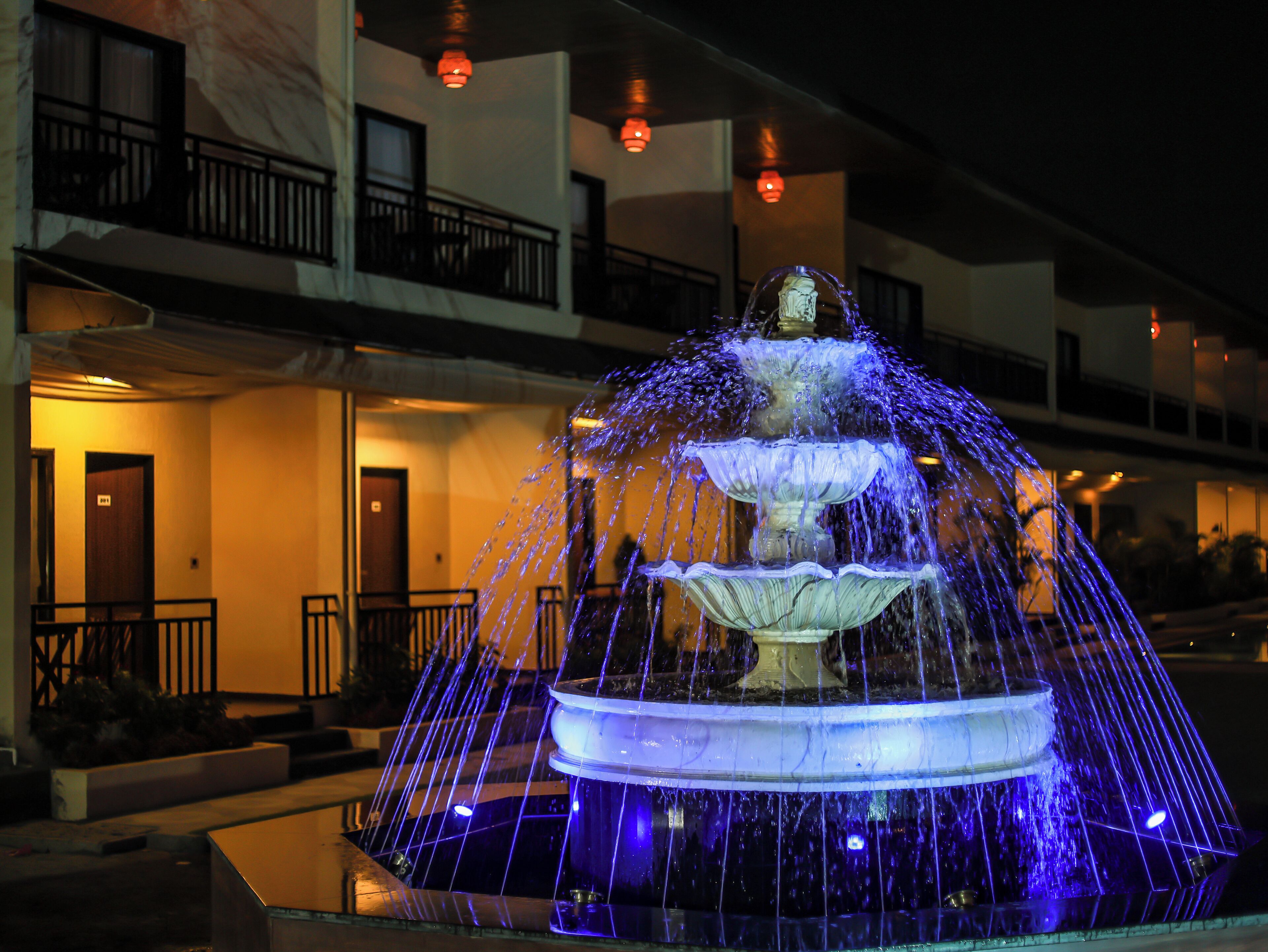 A multi-tiered fountain with water jets, illuminated by blue lights, in front of a building with balconies and doors.