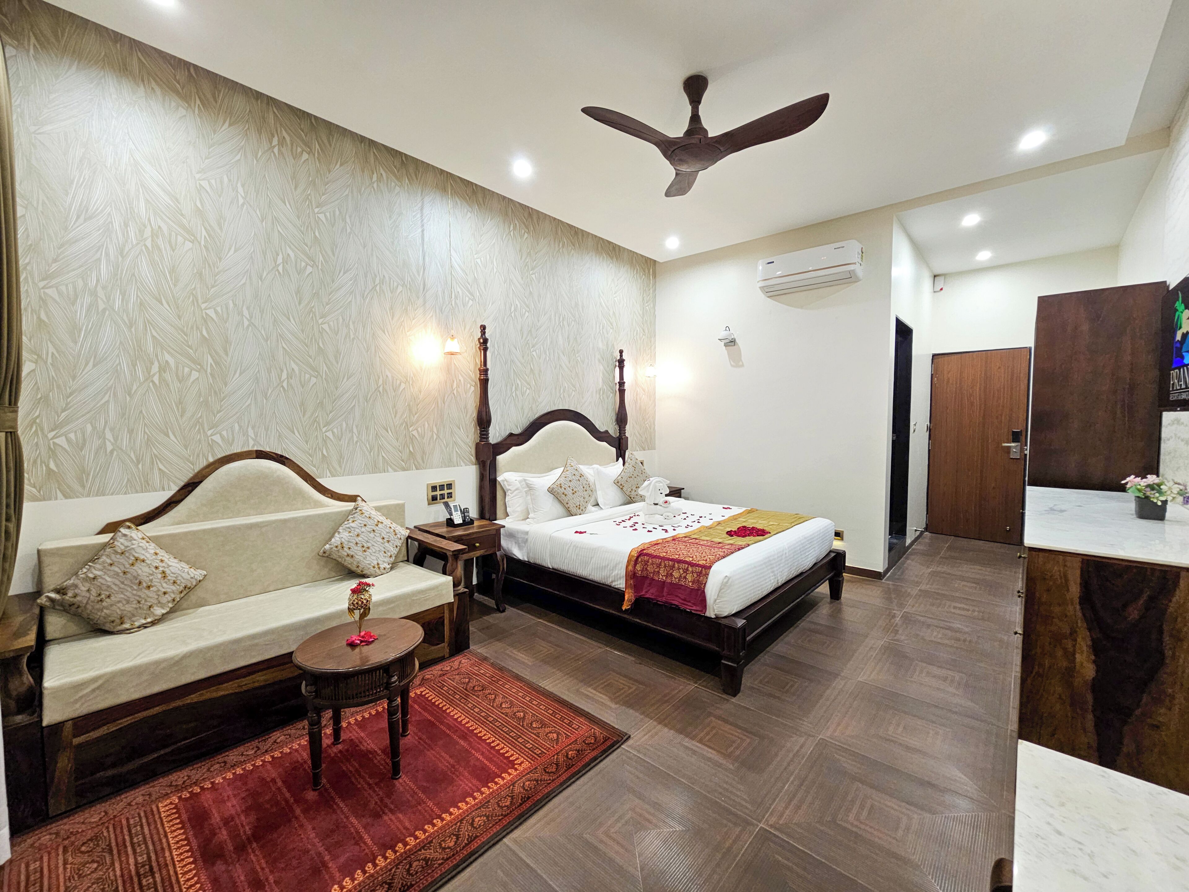 Habitación de hotel con cama, sofá, una mesita, ventilador de techo, aire acondicionado y una puerta de madera.