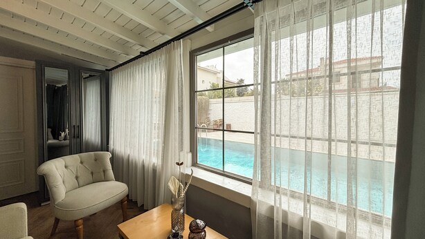 Quarto com vista para a piscina, uma cadeira, uma mesa com objetos decorativos e uma janela com cortinas leves.