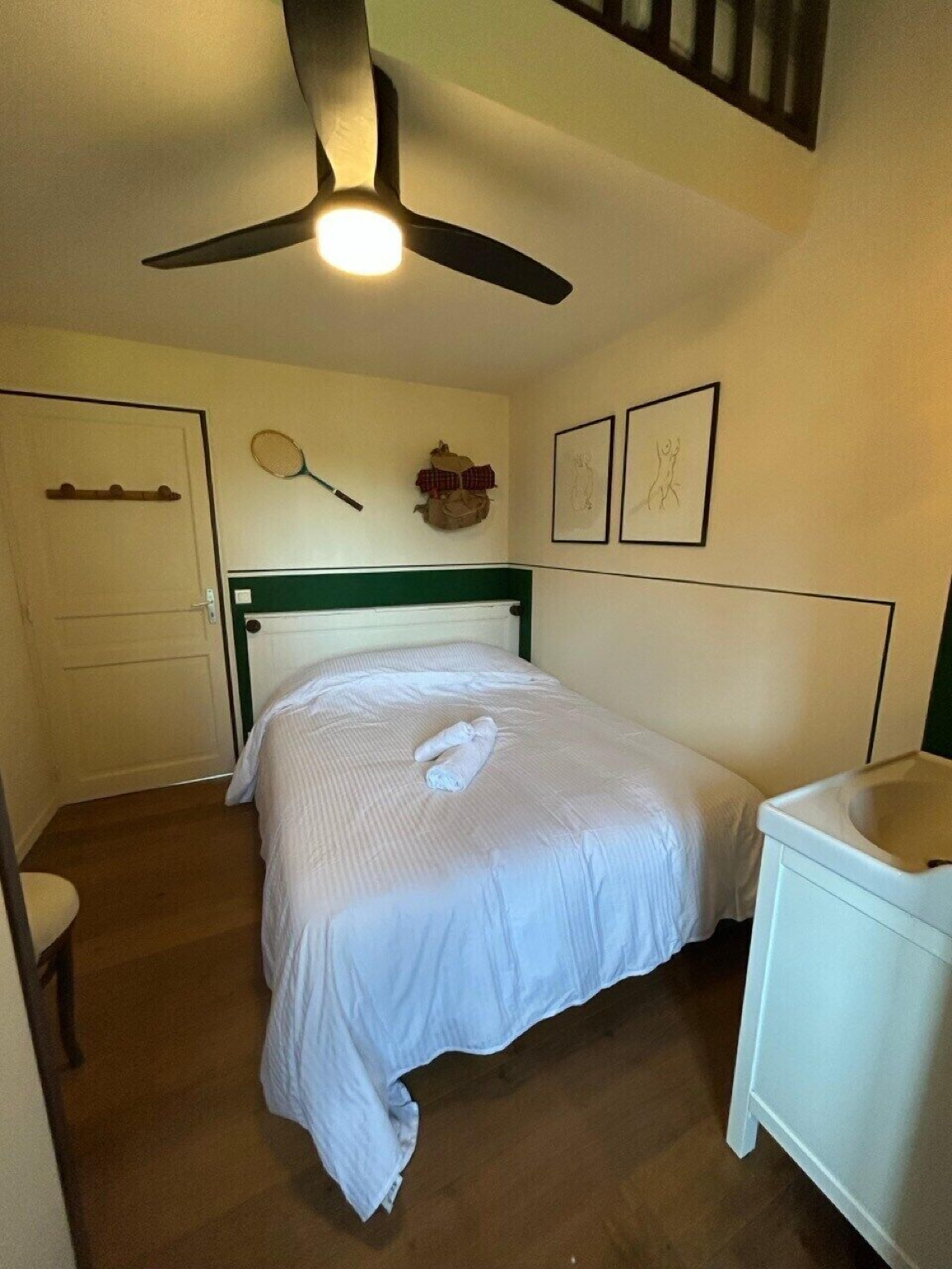 Un dormitorio con cama, ventilador de techo, mesita de noche, inodoro empotrado y un mueble pequeño.