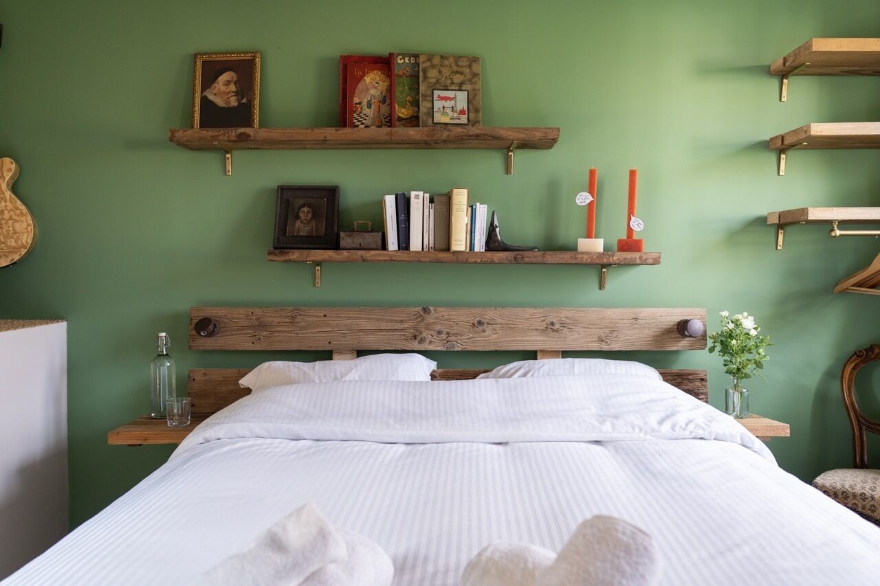A neatly made bed with white linens, wooden headboard, and a green wall with shelves holding books and decorative items.