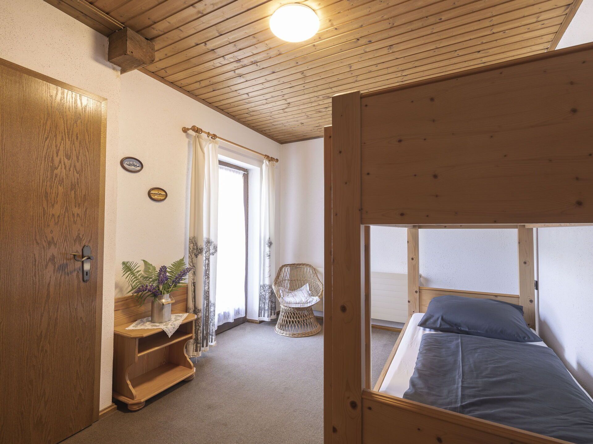 A bedroom with a wooden bunk bed, a wooden desk with a plant, a wicker chair, and a door with a handle.