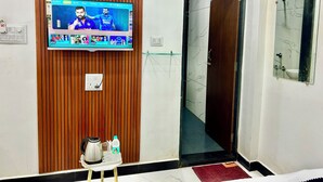 A hotel room with a television mounted on a wooden panel wall, a small table with a tray of tea and snacks, a mirror on the wall, and a door leading to another room. - Hotel Shree (Ujjain)