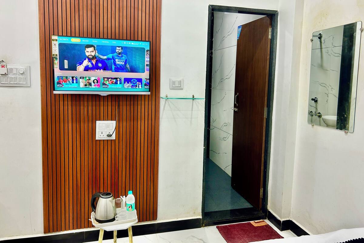 A hotel room with a television mounted on a wooden panel wall, a small table with a tray of tea and snacks, a mirror on the wall, and a door leading to another room.