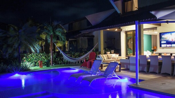 A swimming pool with blue lighting, lounge chairs, a hammock, and a dining area with tables and chairs. - Malawi Resort (Ipojuca)