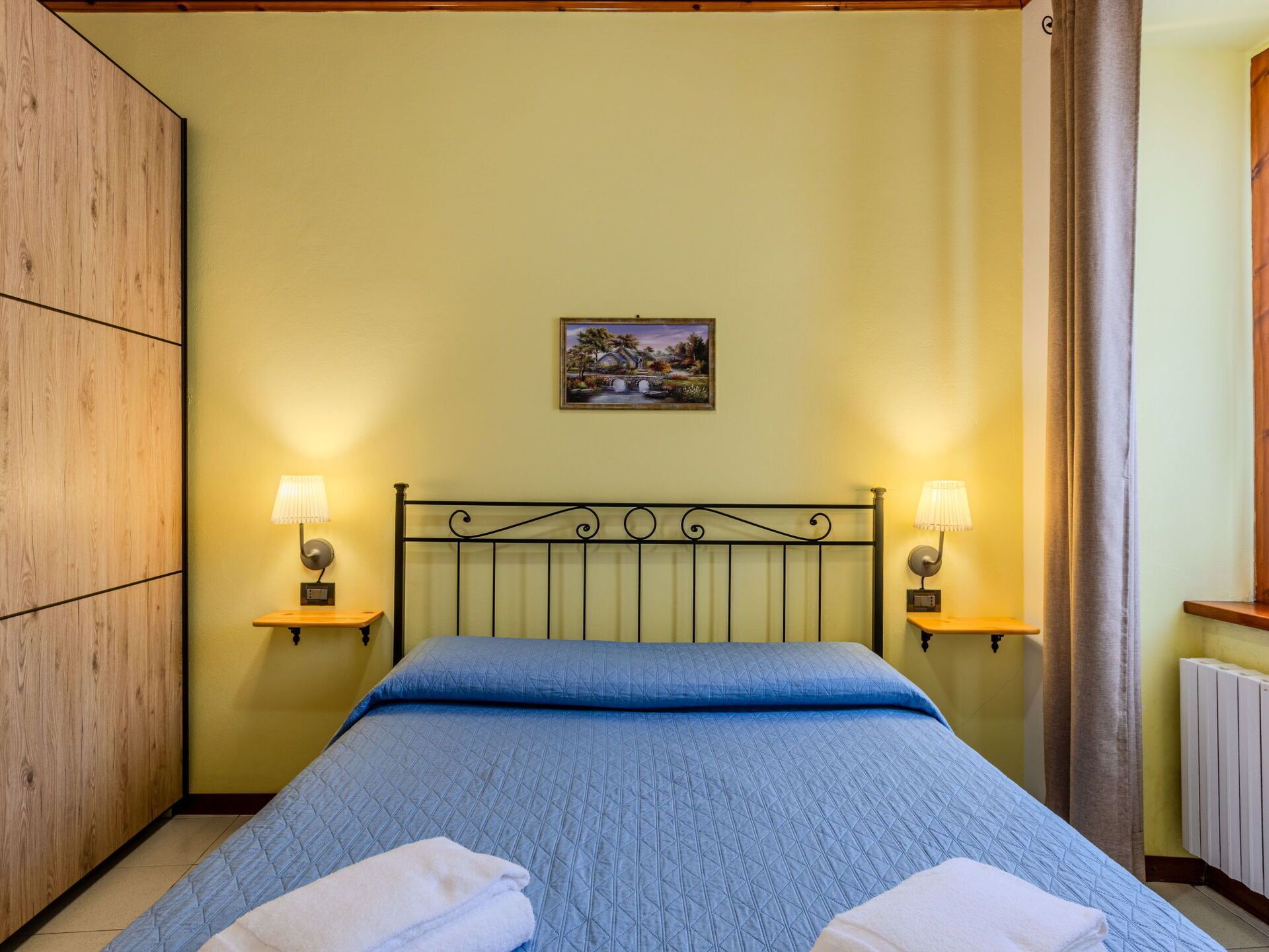 A bedroom with a bed, two bedside lamps, a painting on the wall, and a wooden wardrobe.