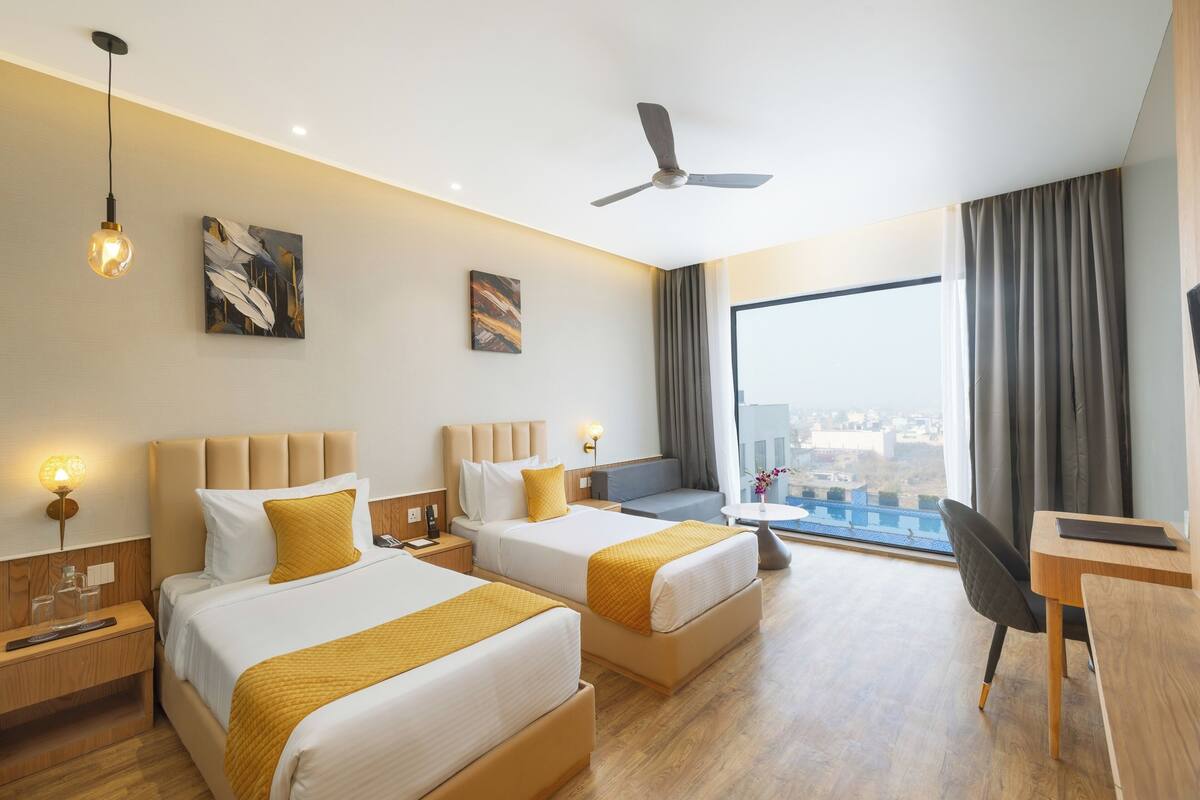 A hotel room with two beds, a desk, a chair, a ceiling fan, and a large window with a city view.