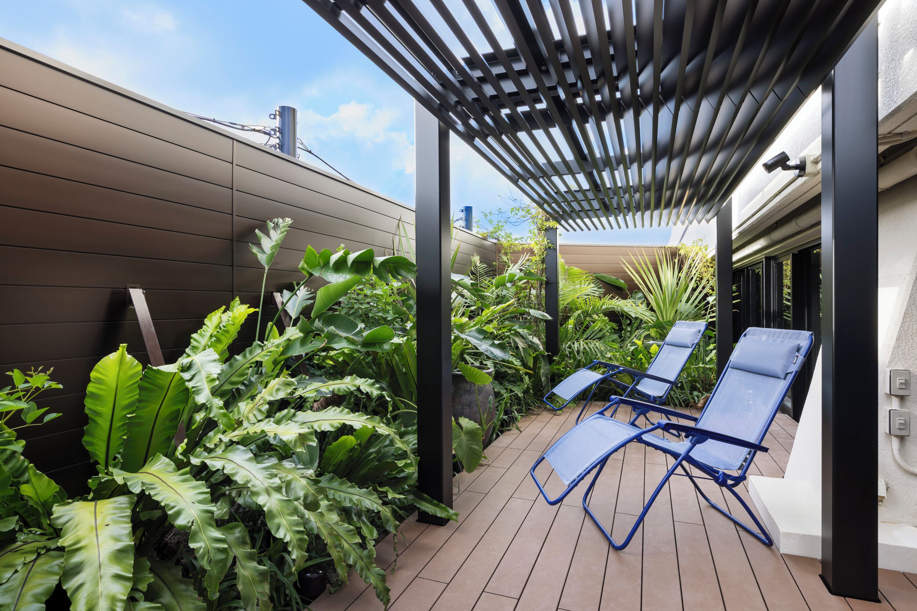 Ein überdachter Außenbereich mit einer Pergola, blauen Lounge-Stühlen und üppigem Grün.