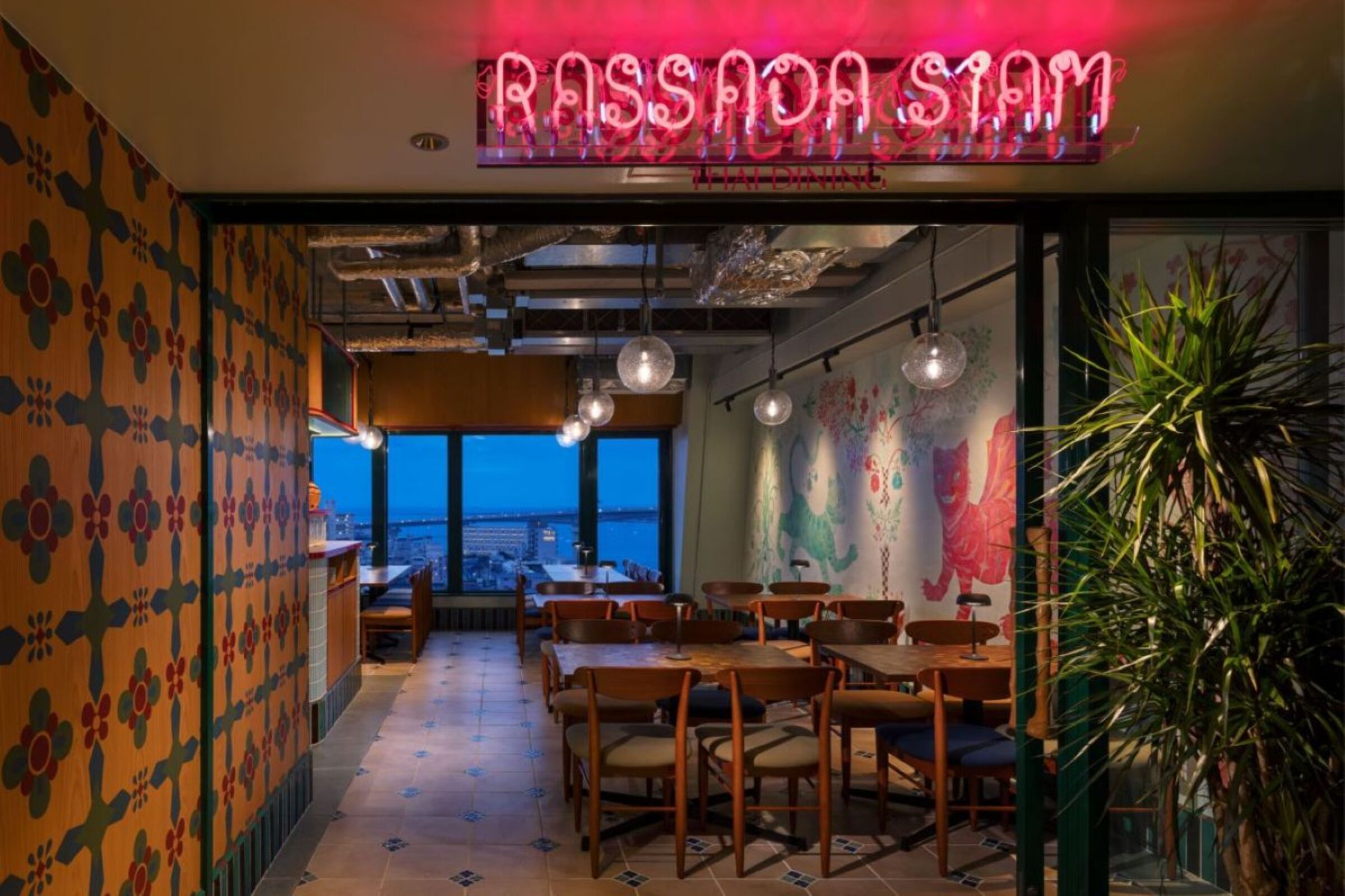 An interior view of a restaurant with a neon sign, patterned walls, and a view of the cityscape.