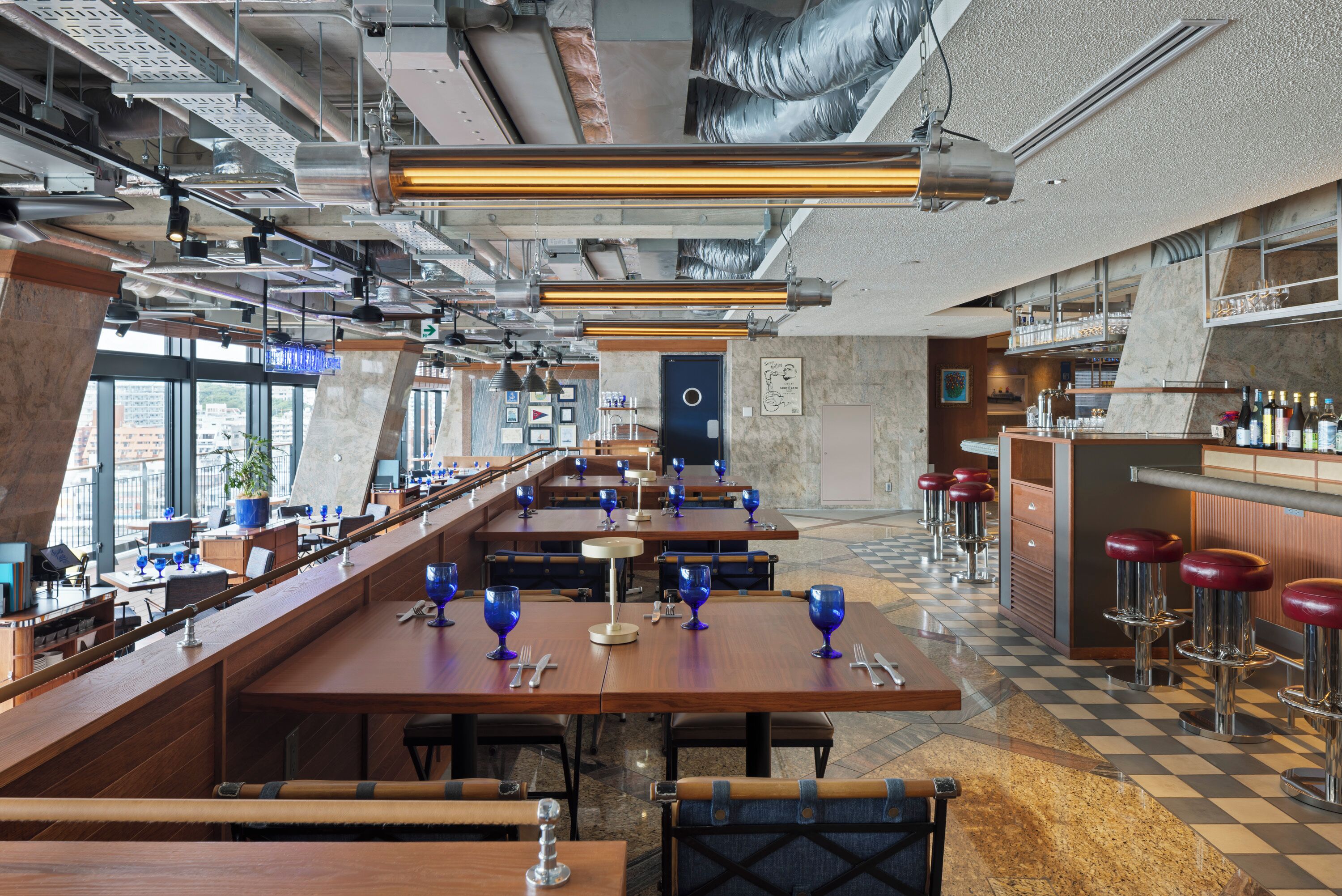 A modern restaurant with high ceilings, exposed ductwork, and a checkered floor. Tables are set with blue glassware and silverware.