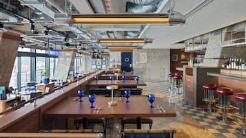 A modern restaurant with high ceilings, exposed ductwork, and a checkered floor. Tables are set with blue glassware and silverware.