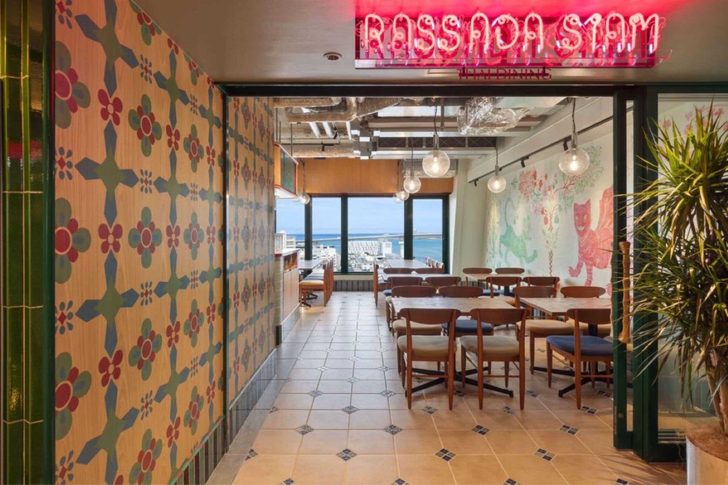 A restaurant interior with a view of the ocean, patterned walls, and hanging pendant lights.