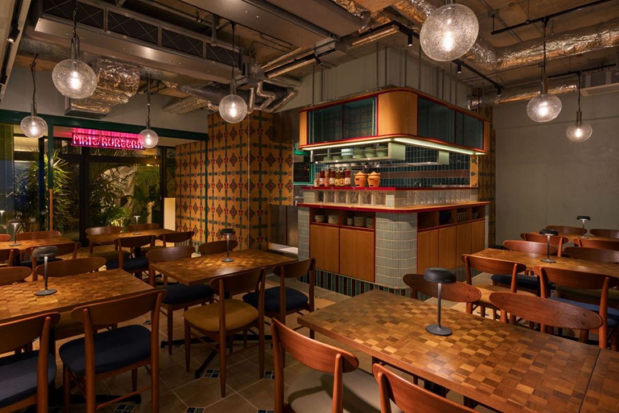 A restaurant interior with wooden tables and chairs, hanging pendant lights, and a kitchen area with shelves and jars.