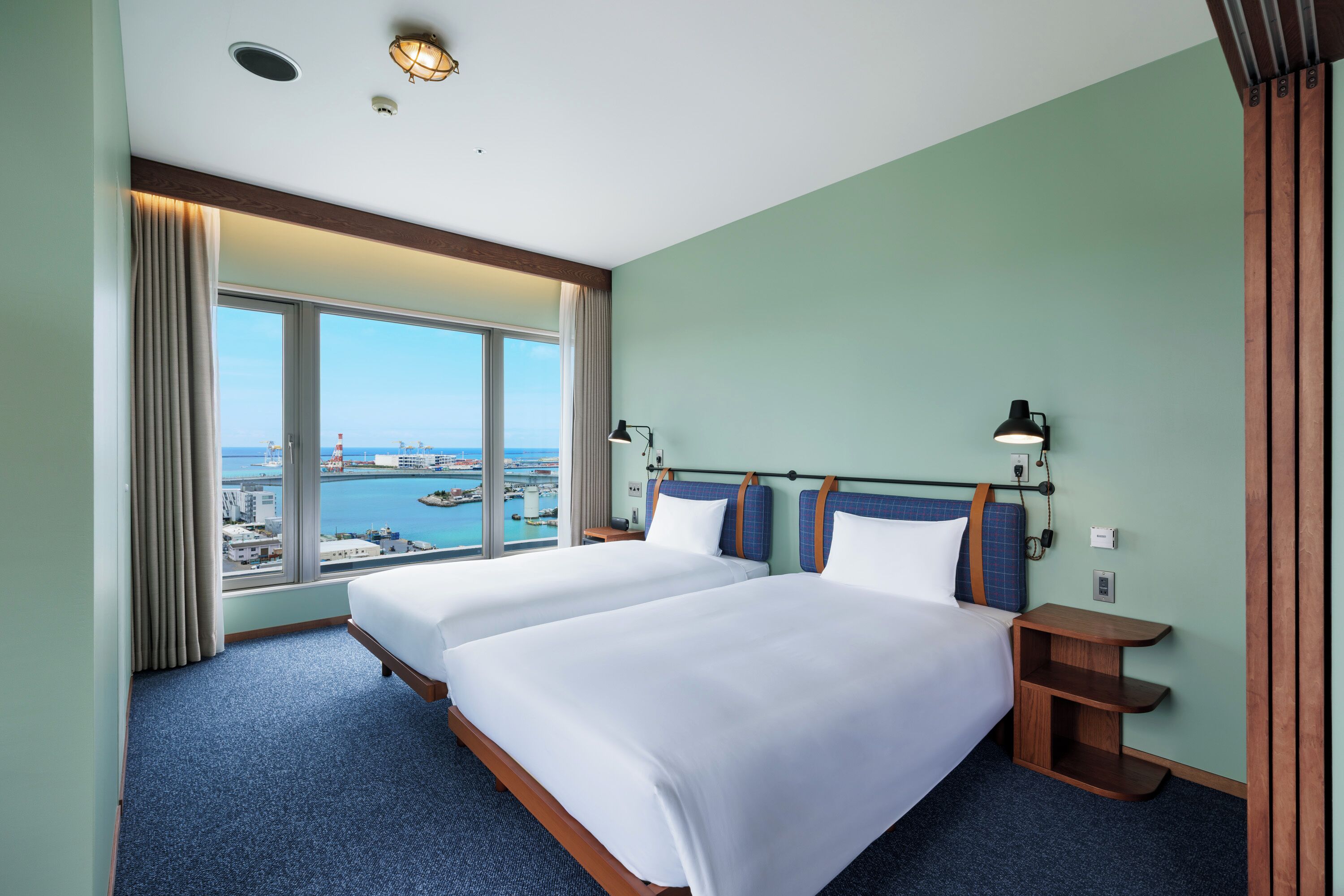 Une chambre d’hôtel avec deux lits, une grande fenêtre donnant sur la mer et les bâtiments environnants, et une table de chevet en bois.