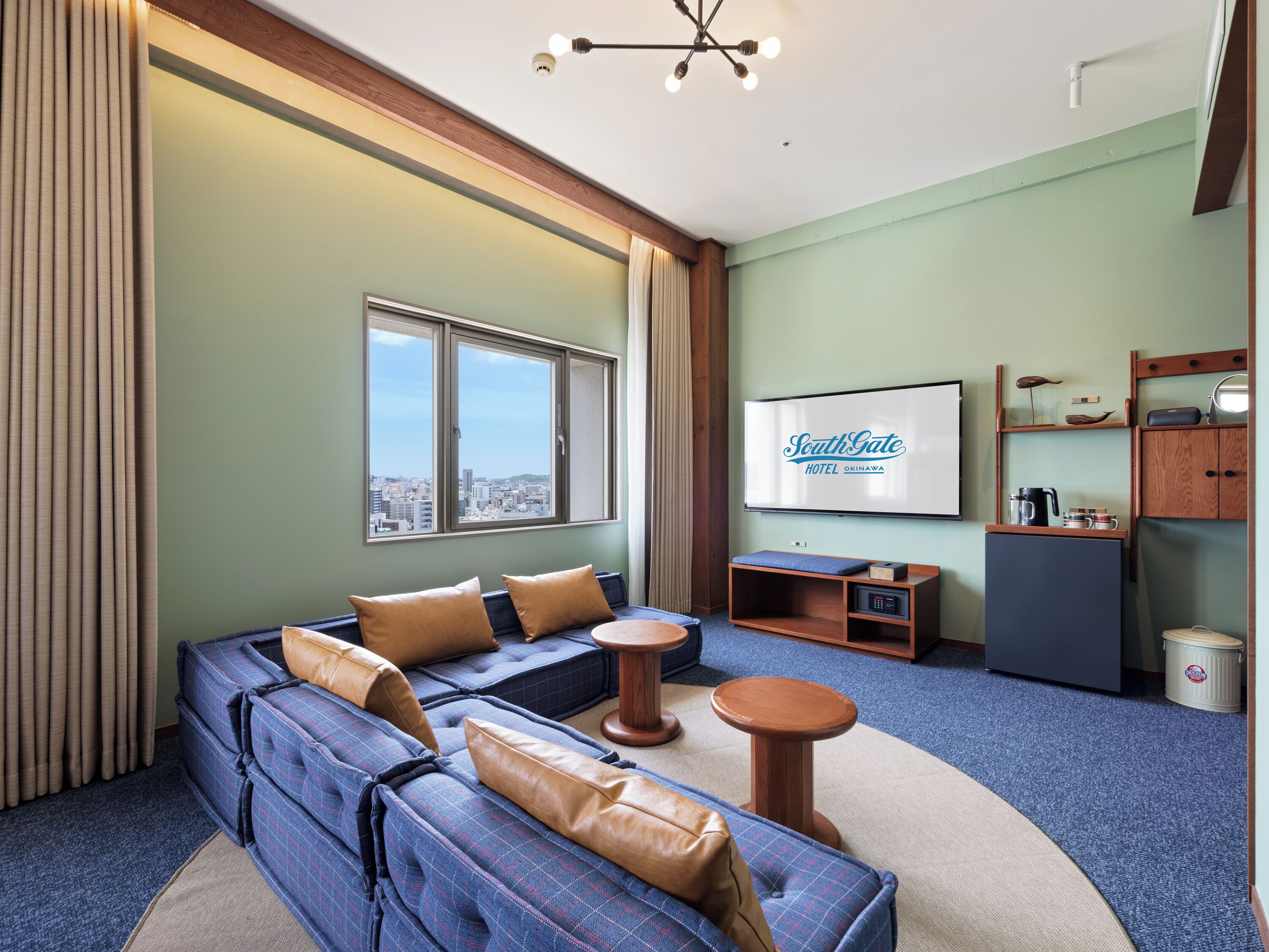 A hotel room with a large window, a sofa, a small table, a TV, and a kitchenette.