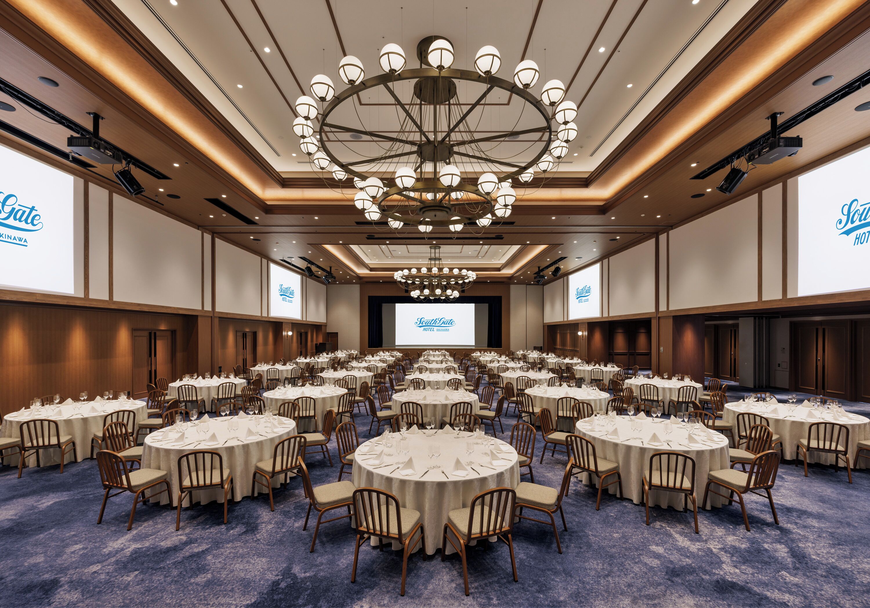 A large conference room with multiple round tables, a central chandelier, and a projection screen at the front.