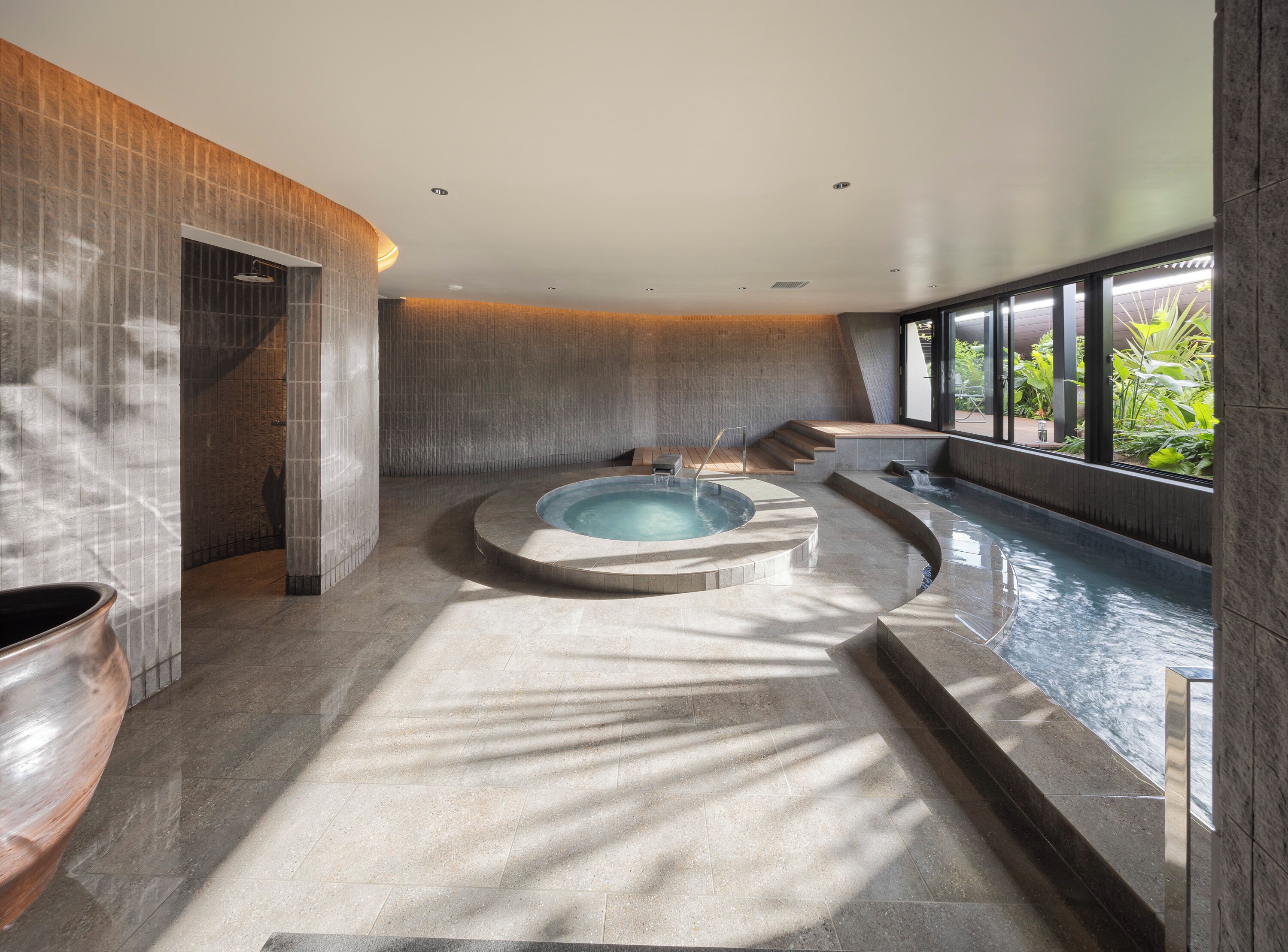 A modern spa with a central hot tub, stone walls, and a view of greenery.