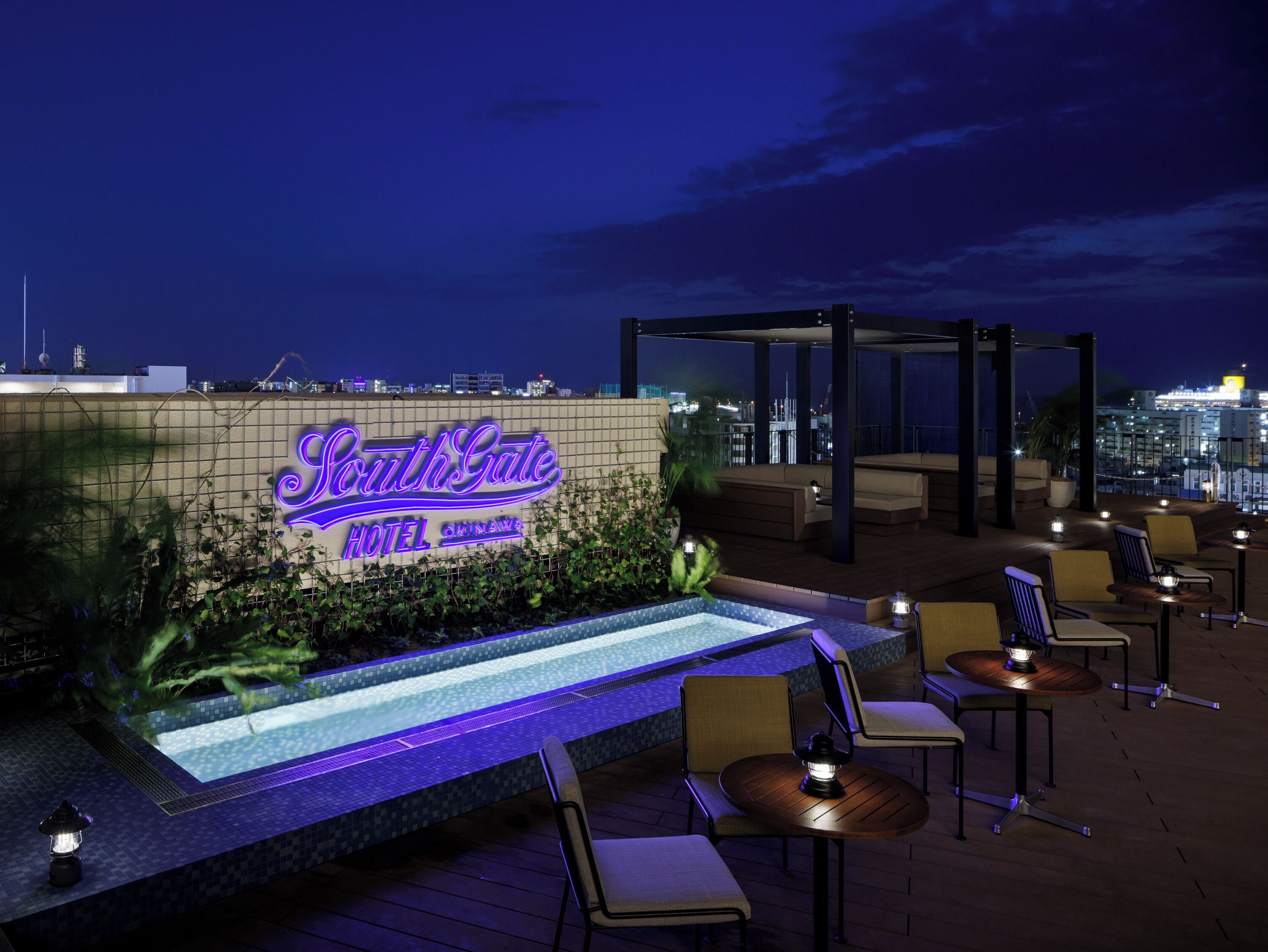 A hotel sign, outdoor seating area, and a pool with a city view at night.