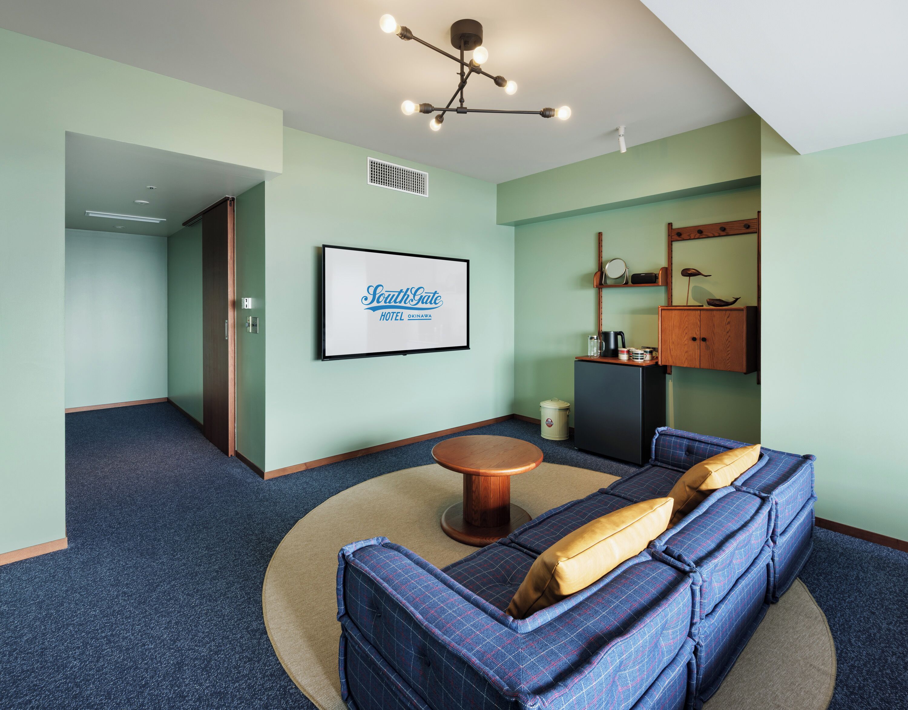 Habitación de hotel con un sofá azul, una mesita de centro redonda, una lámpara en el techo y un cartel enmarcado en la pared.