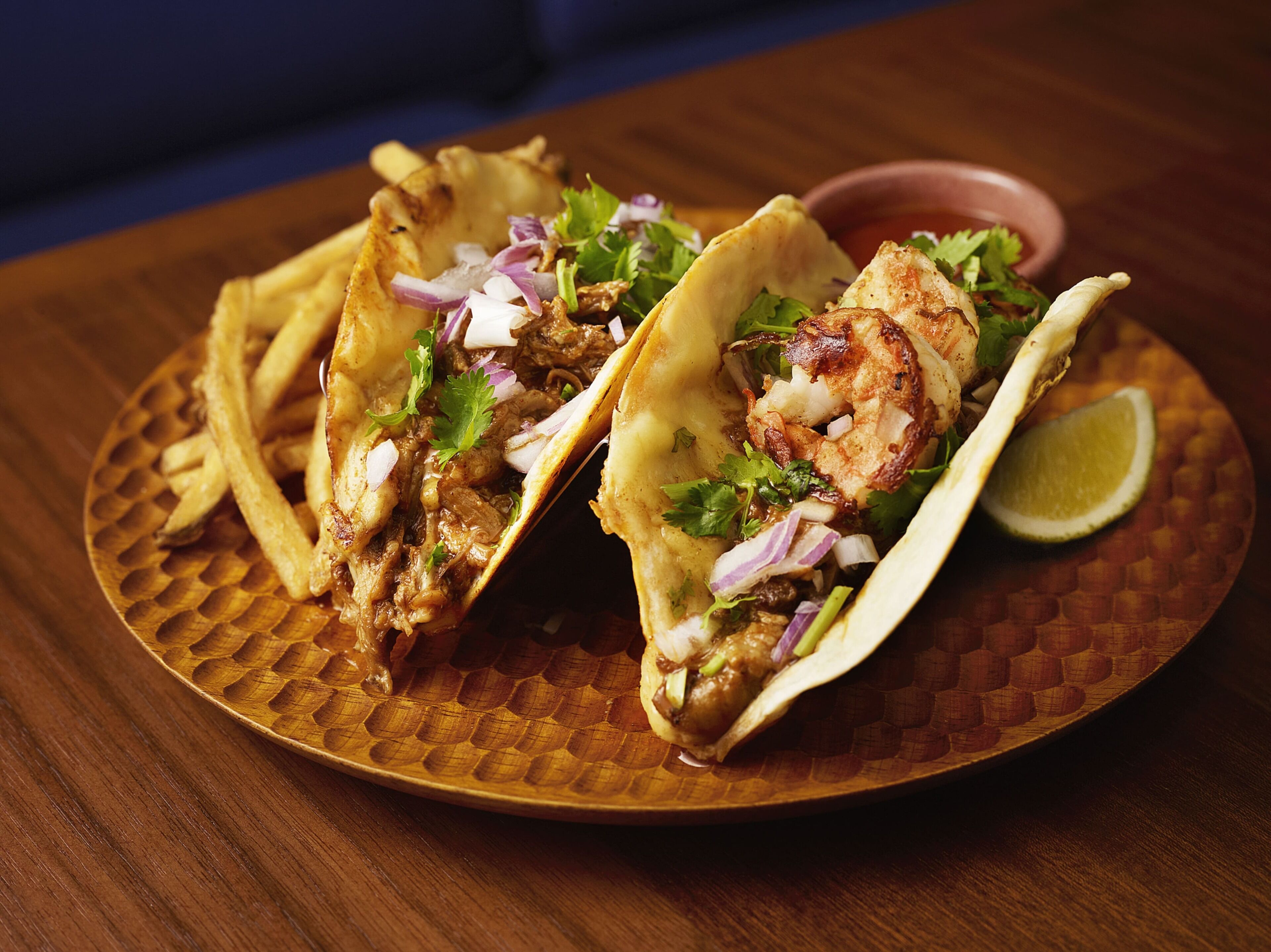 Two tacos with meat, onions, and cilantro on a wooden plate with fries and a lime wedge.