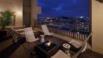 A balcony with a fire pit, lounge chairs, and a view of the cityscape at night.