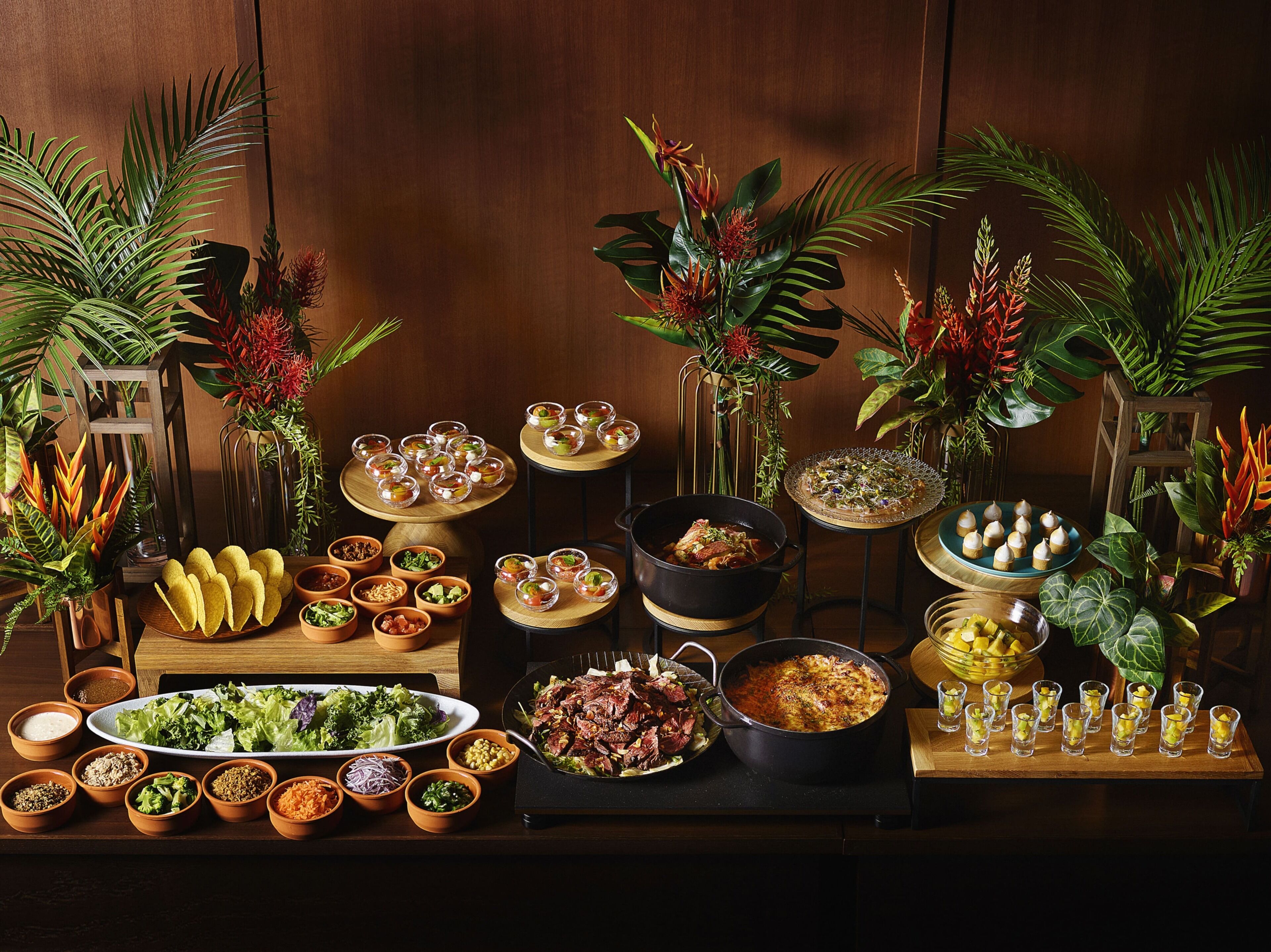 A variety of dishes and drinks on a wooden table, surrounded by decorative plants.